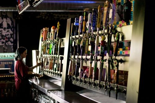 A bartender stands at a bar with numerous colorful beer taps in a dimly lit setting.