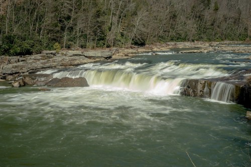 A serene river scene featuring a waterfall cascading over rocky ledges, surrounded by trees and a calm greenish water.