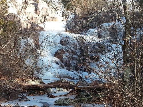 A frozen waterfall cascading over rocks, surrounded by bare trees and winter scenery.