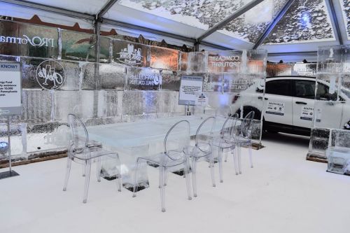 A transparent ice dining table and chairs inside a tent, with ice walls and a vehicle in the background.
