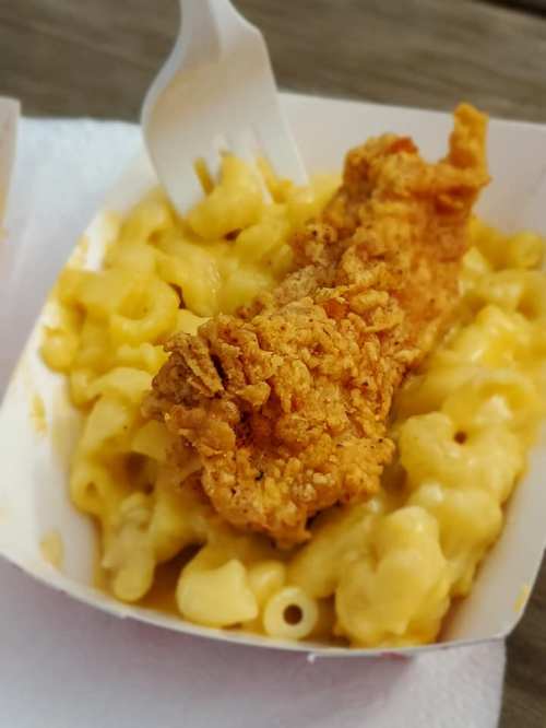 A serving of macaroni and cheese topped with a crispy fried chicken strip in a takeout container.