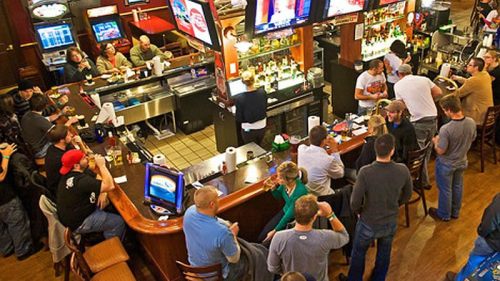 A busy bar scene with patrons socializing, drinks on the counter, and multiple TVs showing sports.