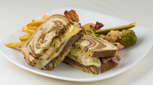 A close-up of a sandwich with layers of meat, cheese, and sauerkraut, served with fries and assorted vegetables on a plate.