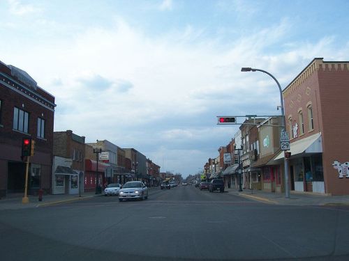 A quiet downtown street with shops on either side and a traffic light showing red under a cloudy sky.