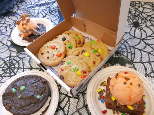 A box of colorful cookies with chocolate chips and two decorated desserts on plates, set on a spiderweb tablecloth.