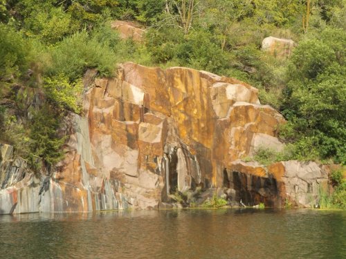A rocky cliff with colorful layers reflects in calm water, surrounded by lush greenery.