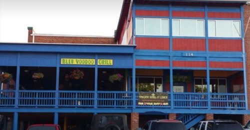 Colorful exterior of Bleu Voodoo Grill, featuring blue and red walls, large windows, and hanging flower baskets.