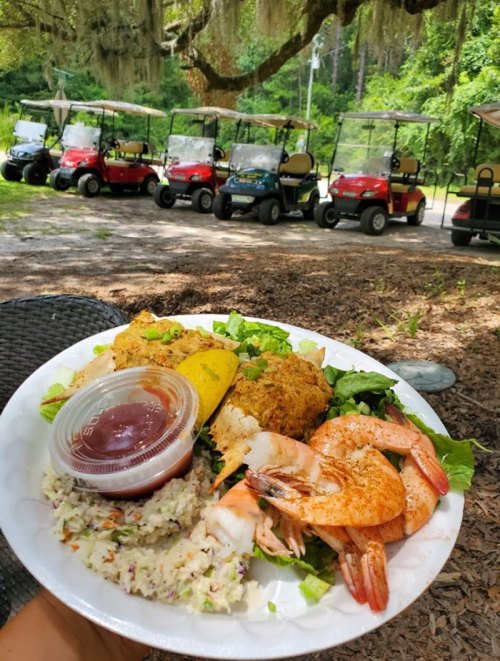 A plate of shrimp, coleslaw, and cornbread, with golf carts parked under trees in the background.