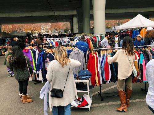 A bustling outdoor market with people browsing clothing racks under a highway overpass. Tents are set up in the background.