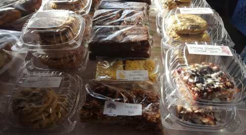 A variety of baked goods in clear containers, including cookies, brownies, and pecan pie bars, displayed on a table.