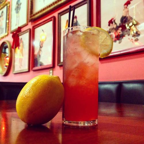 A tall glass of colorful drink with ice and lemon slices, next to a whole lemon, set against a pink wall with framed art.
