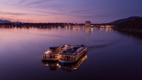 Two illuminated boats on a calm lake at dusk, with a scenic shoreline and city lights in the background.
