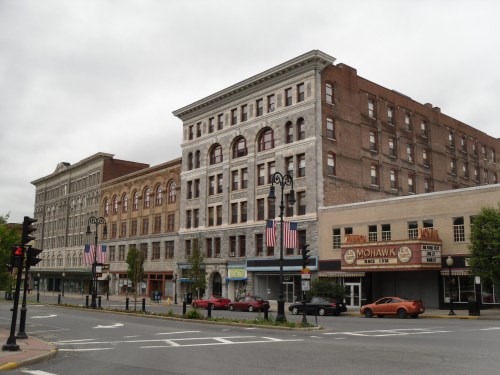 Historic buildings line a street, featuring various architectural styles and American flags, under a cloudy sky.