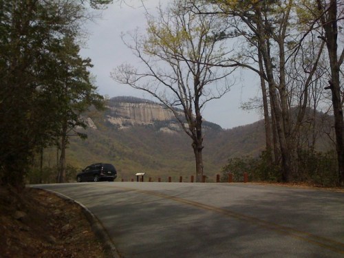 A winding road curves through a forested area, with a rocky mountain in the background and a vehicle parked nearby.