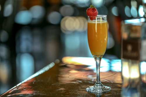 A sparkling cocktail in a tall glass, garnished with a strawberry, sits on a reflective table.