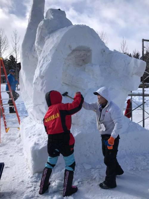Two people sculpt a large snow statue, with one using a tool and the other assisting, surrounded by snow and trees.