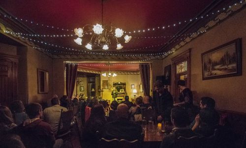 A cozy room filled with people, dimly lit, with string lights and a chandelier, focused on a performer on stage.