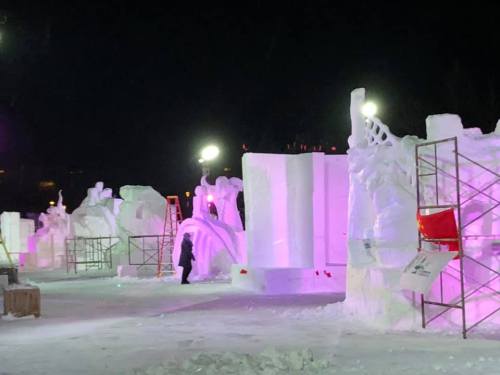 Snow sculptures illuminated at night, with artists working and scaffolding in a winter landscape.
