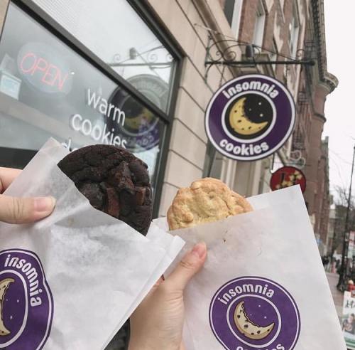 Two hands hold cookies wrapped in paper outside an Insomnia Cookies shop, with a sign in the background.
