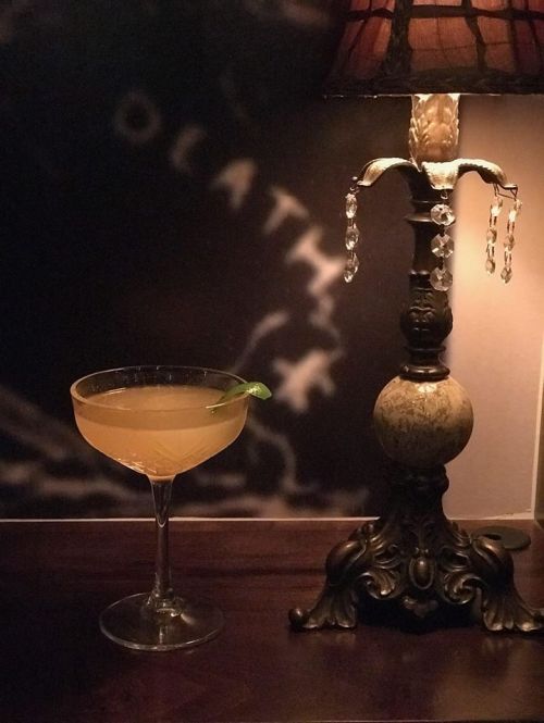 A cocktail in a coupe glass sits on a wooden table beside an ornate lamp with a stained glass shade.