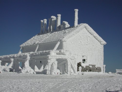 A snow-covered house with thick layers of snow on the roof and walls, set against a clear blue sky.