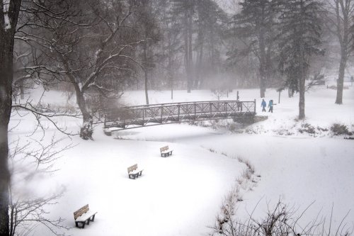A snowy landscape with a bridge and two figures walking in the distance, surrounded by trees and benches.