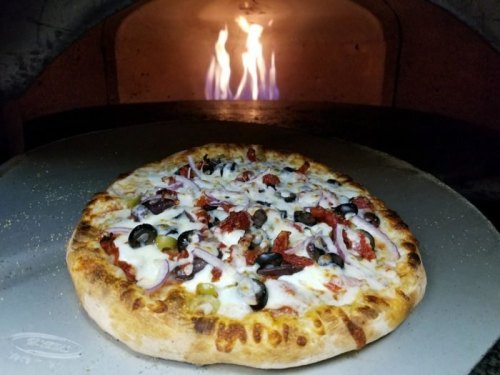A freshly baked pizza with olives, onions, and sun-dried tomatoes, placed in a wood-fired oven with flames in the background.