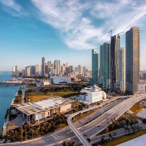 Aerial view of a vibrant city skyline with modern skyscrapers, waterfront, and a clear blue sky.