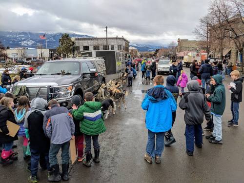 A crowd of children gathers on a street, observing a truck and a display of logs, with mountains in the background.