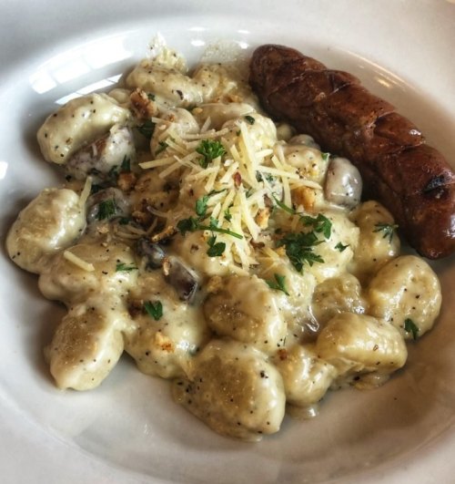 A plate of creamy gnocchi topped with cheese and herbs, accompanied by a grilled sausage.