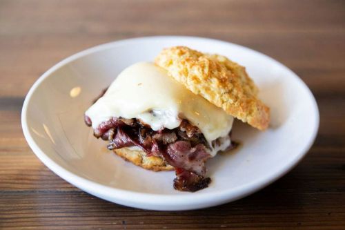 A savory sandwich with tender meat and melted cheese, served on a flaky biscuit, on a white plate.