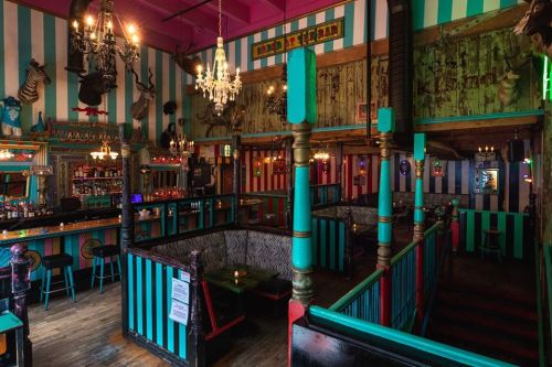 Colorful bar interior with striped walls, chandeliers, and eclectic decor, featuring cozy seating areas and a vibrant atmosphere.