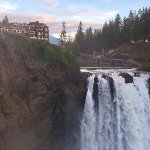 A waterfall cascades down a cliff beside a large building, surrounded by trees and a cloudy sky.