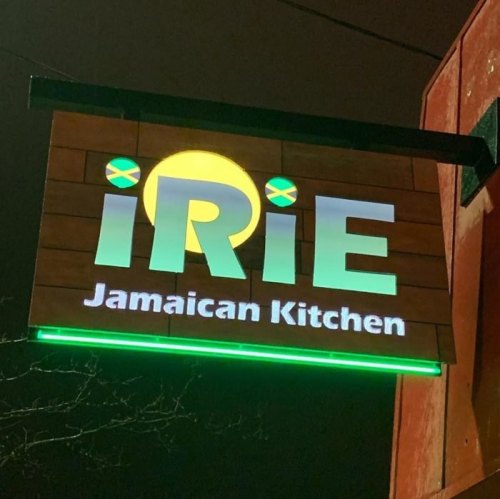 Sign for "iRiE Jamaican Kitchen" illuminated at night, featuring Jamaican flag colors and a circular logo.