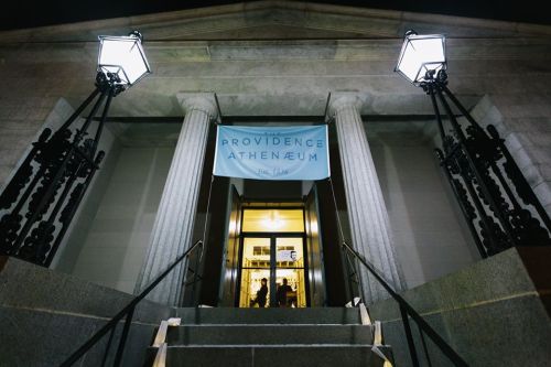 Entrance of the Providence Athenaeum at night, featuring columns, lamps, and a large banner.