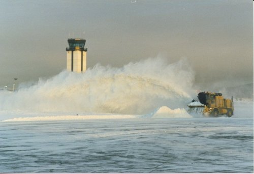 A snowplow clears snow at an airport, with a control tower visible in the background under a cloudy sky.