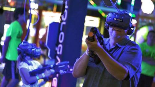 A person and a child wearing VR headsets, aiming with toy guns in a colorful arcade setting.