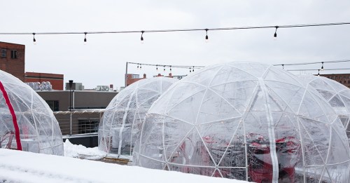 Clear igloos on a rooftop, surrounded by snow, with string lights overhead and a cloudy sky in the background.