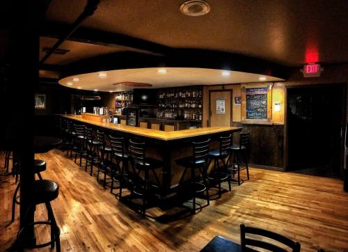 A dimly lit bar interior with a long wooden counter and empty stools, featuring shelves of drinks in the background.