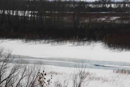 A snowy landscape with a winding river, surrounded by bare trees and patches of grass.