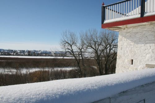 A snowy landscape with a stone building and a clear blue sky, overlooking a river and distant hills.