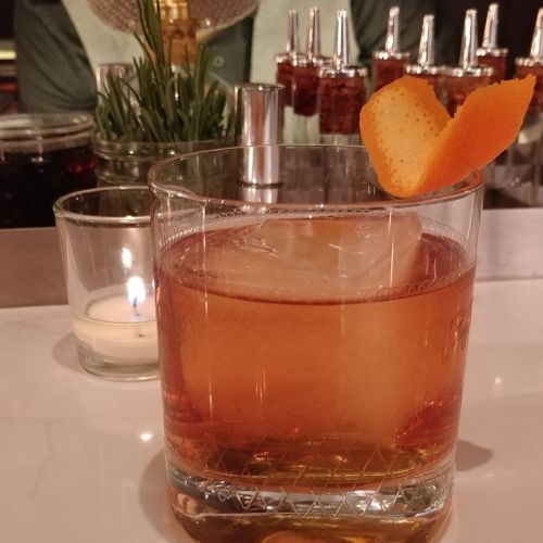A cocktail in a glass with ice, garnished with orange peel, sits on a bar next to a small candle.
