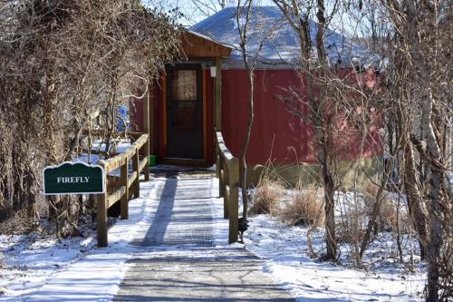 A wooden walkway leads to a red yurt surrounded by trees, with a sign reading "Firefly" and snow on the ground.