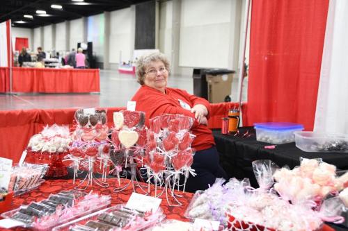 A smiling vendor in a red shirt sits behind a table filled with various candies and treats at a fair.