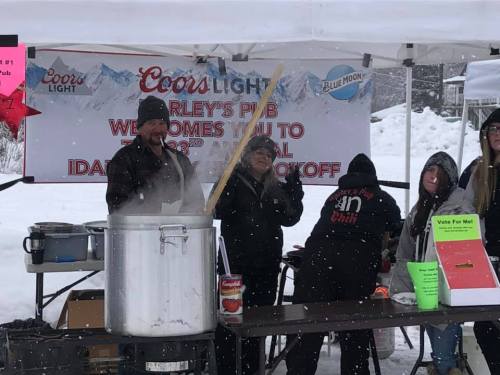 A group of people at a chili cook-off event, with a large pot and a banner welcoming attendees. Snow is visible.