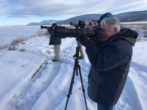 A photographer with a large camera lens stands on snow, capturing images, while another person observes in the background.