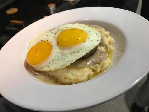 A plate of creamy mashed potatoes topped with sausage, gravy, and two sunny-side-up eggs, garnished with pepper.
