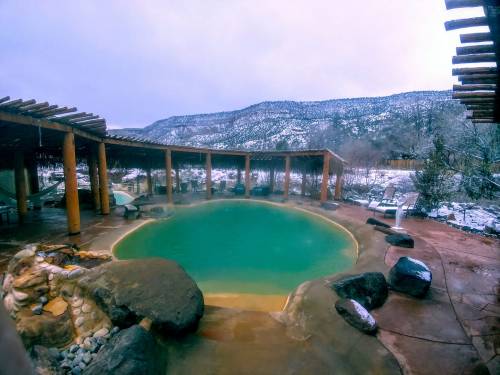 A serene hot spring surrounded by rocks and wooden structures, with snow-covered mountains in the background.