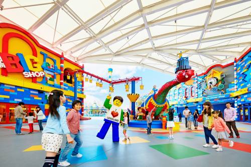 Colorful indoor LEGO shop with children playing, a LEGO dinosaur, and a costumed character in a vibrant, cheerful atmosphere.