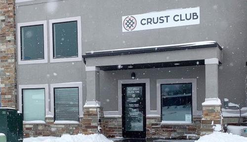 Exterior of Crust Club building with snow on the ground and falling, featuring large windows and a sign above the entrance.